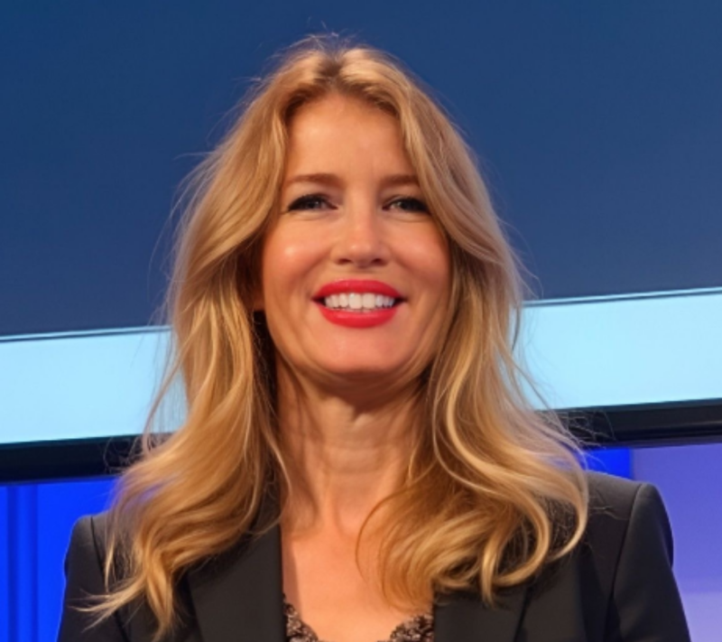 A woman with long blonde hair and a bright smile stands in front of a blue and white background, wearing a black jacket.