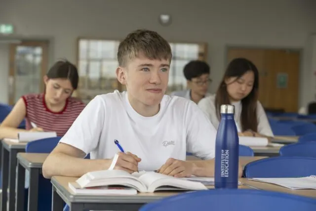 a boy sits, listening, in class. He holds a pen.