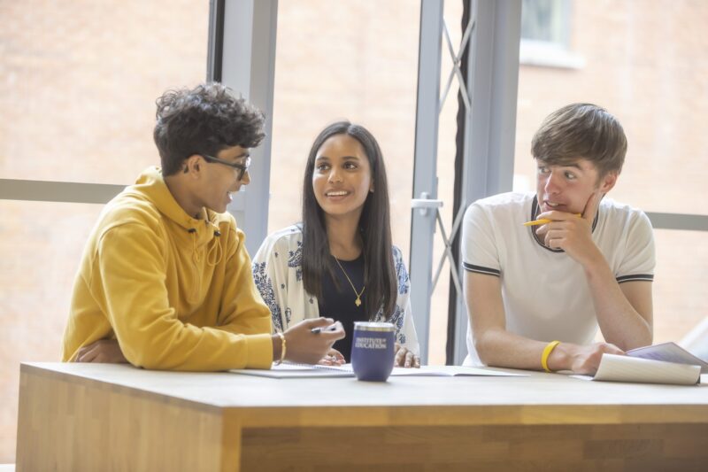 Three Institute of Education students chatting at a table with notebooks and an Institute of Education branded cup, in a bright glass-walled study space.