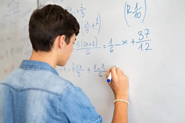 A pupil writes algebraic equations with fractions on a whiteboard using a blue marker.