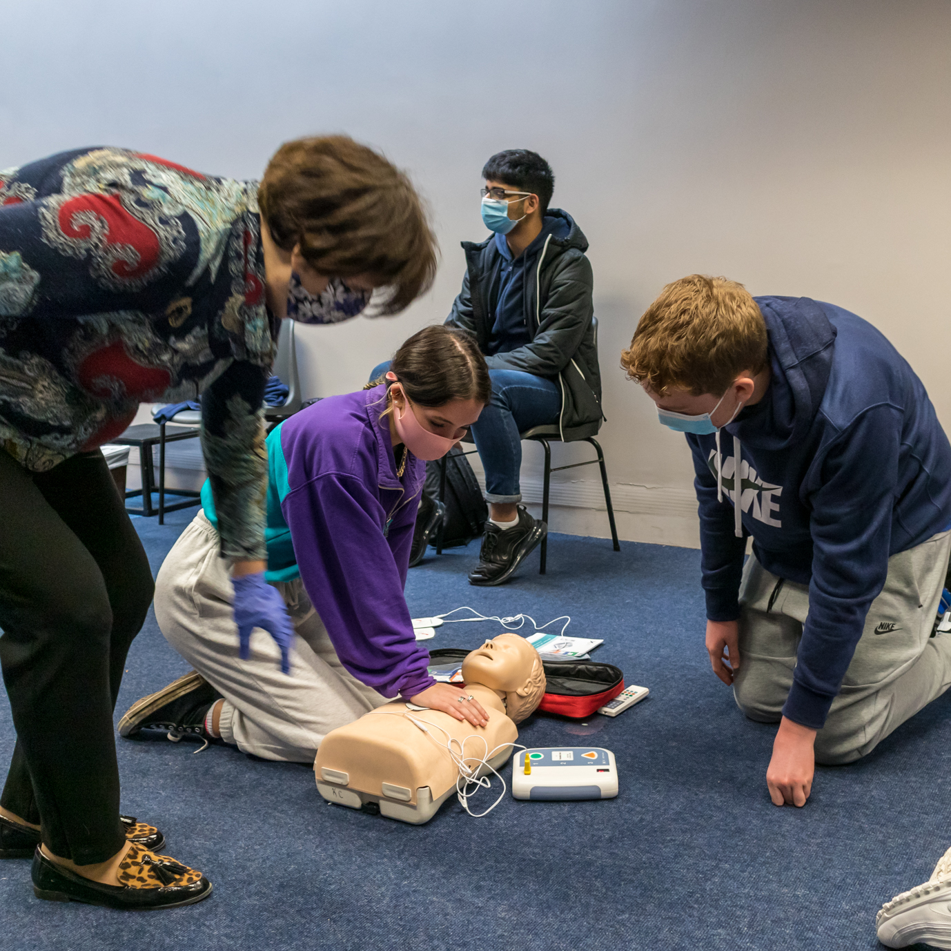 Instructor teaching student CPR, and another person is about to kneel beside them