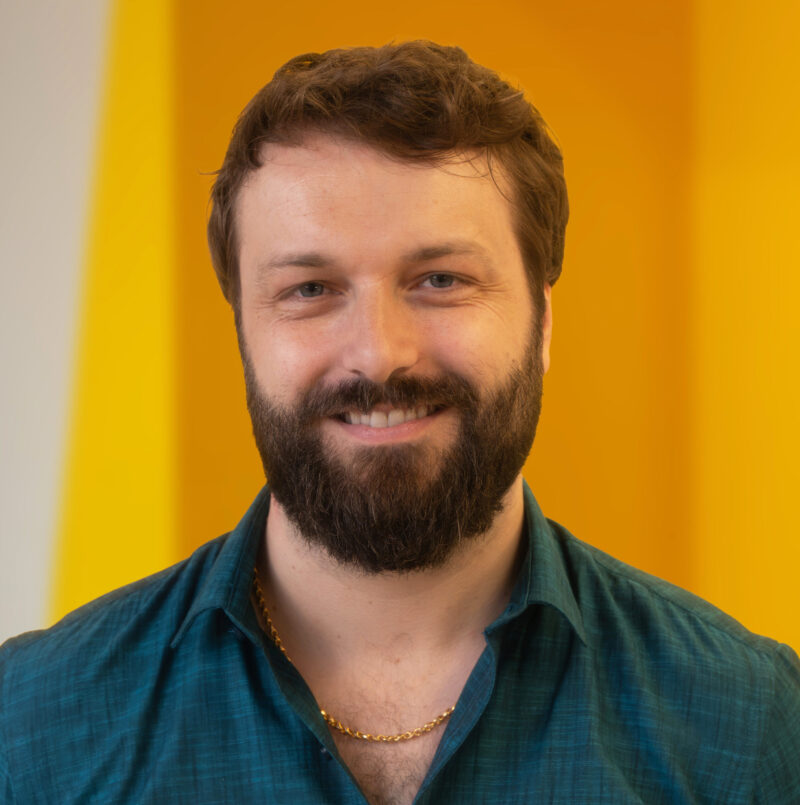 A man with a beard and moustache, wearing a teal shirt and gold chain, smiles at the camera. The background is yellow and white.