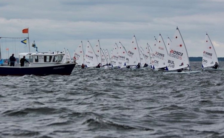 A large number of sailboats, all lined up with a larger fishing boat supervising