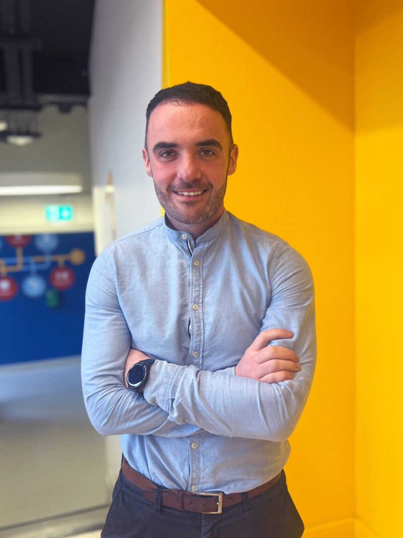 A smiling man with short dark hair wearing a light blue button-up shirt and a black wristwatch stands with his arms folded in front of a yellow wall.