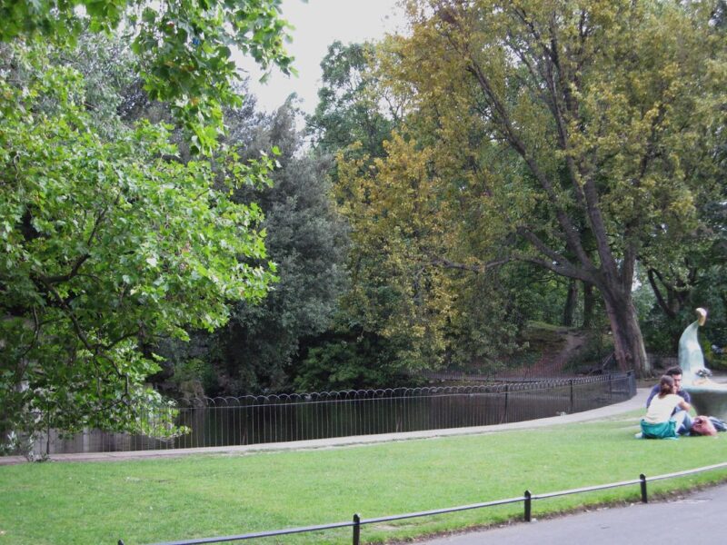 Park scene with green lawns, mature trees and a couple sitting on the grass beside a path (St Stephen's Green style setting).
