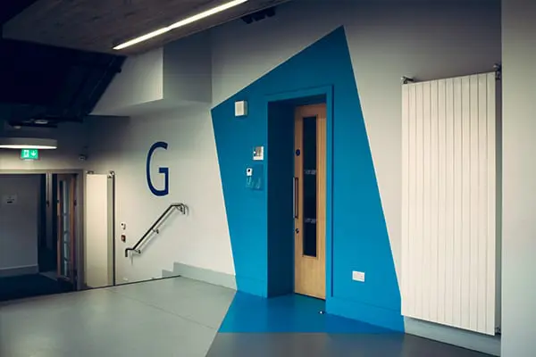 A corridor with a large angular blue accent wall around a wooden door, marked with a bold letter G, next to an exit sign and staircase.
