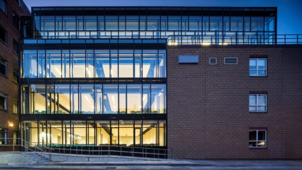 A modern glass extension with lit interior classrooms, joined to an older brick building, photographed at dusk.
