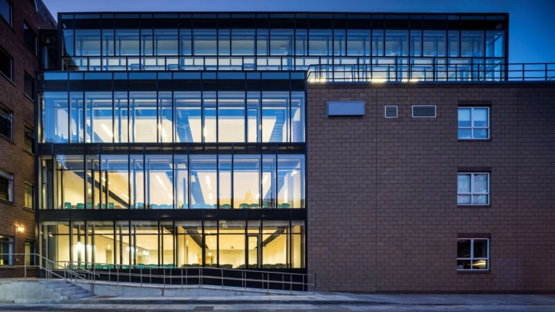 A modern glass extension with lit interior classrooms, joined to an older brick building, photographed at dusk.