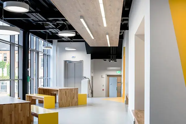 A bright modern corridor with exposed black ceiling, wooden benches and tables, and floor-to-ceiling windows, leading to a lift and doorway.