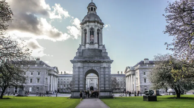 Trinity College Dublin's Campanile bell tower, viewed from the front lawn with students and cyclists nearby.