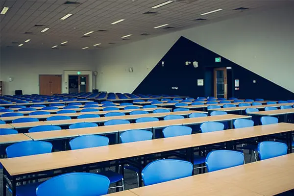 A large empty exam or lecture hall with rows of blue chairs and long tables, and a bold triangular dark accent wall at the front.
