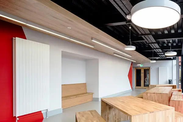 A bright corridor with wooden bench seating, a red angular accent wall, wooden tables, and an exposed black ceiling with round pendant lights.
