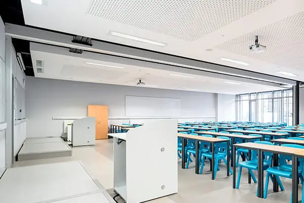 An empty classroom with rows of blue desks and chairs, a lectern, and a wall-mounted screen at the front.
