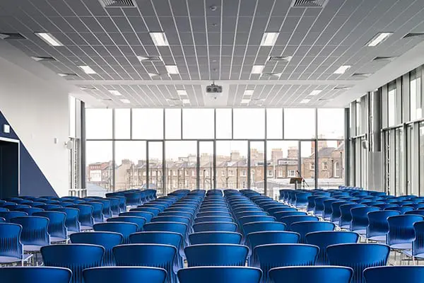 A large empty lecture hall with rows of blue chairs facing a projector screen, floor-to-ceiling windows showing Dublin rooftops beyond.
