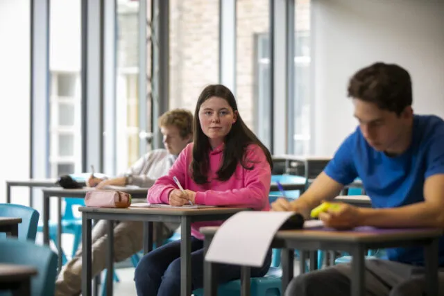 Three students sit at desks in a brightly lit classroom. A girl in a pink hoodie looks forward whilst writing. Two boys, one in a blue shirt and one in white, are focused on their work.