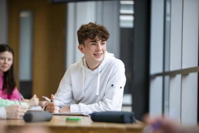 A young person with curly brown hair wearing a white hoodie smiles whilst sitting at a desk with notebooks and pens, looking to the side in a brightly lit room.