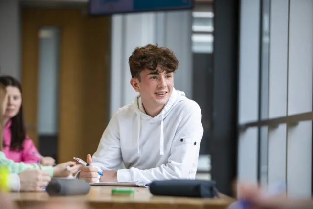 A young person in a white hoodie sits at a table, smiling and holding a pen, with notebooks and other people nearby in a bright, modern classroom.