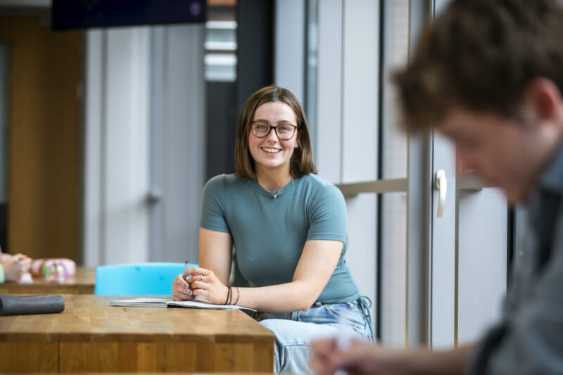 Female student smiling at a desk with a notebook and blue folder, seated by a window.