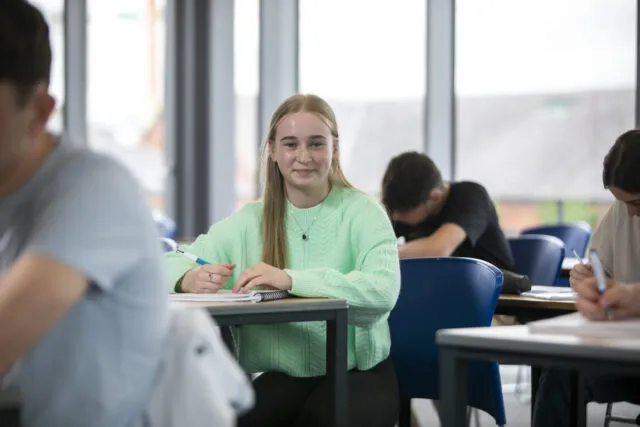 A student in a green jumper sits at a desk, smiling and writing in a notebook, whilst others around her focus on their work in a bright classroom.