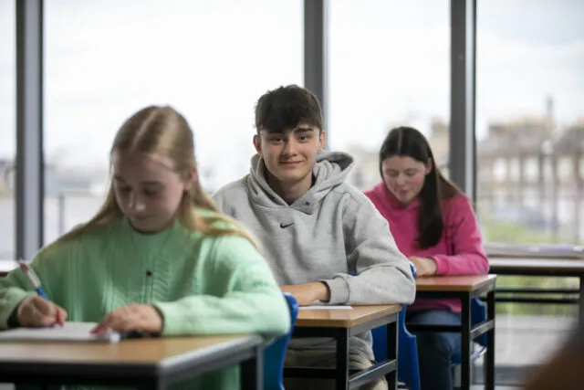 Three students sit at desks in a classroom, writing on paper. The boy in the middle, wearing a grey hoodie, looks up and smiles at the camera, whilst the two girls beside him focus on their work. Large windows are in the background.