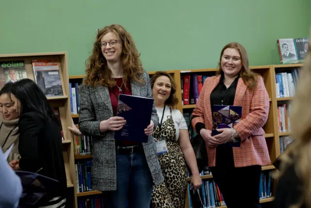 Two women smiling at an awards or presentation event, standing in front of bookshelves with a third woman partly visible.