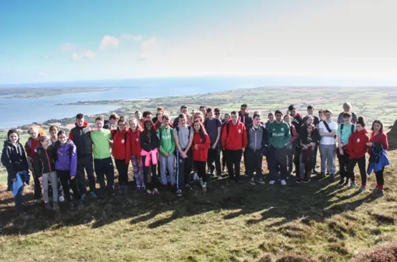 A large group of students posing together on a hilltop overlooking the sea, on a school trip.