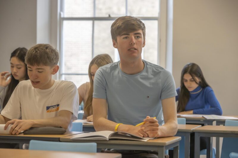 A student sitting at a desk in class, with classmates writing in notebooks around him.