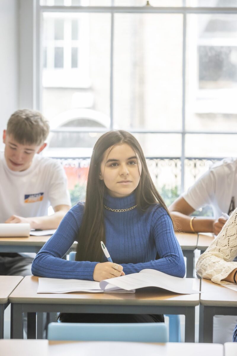 A student sitting at a desk in class, looking towards the camera, with classmates writing in notebooks around her.