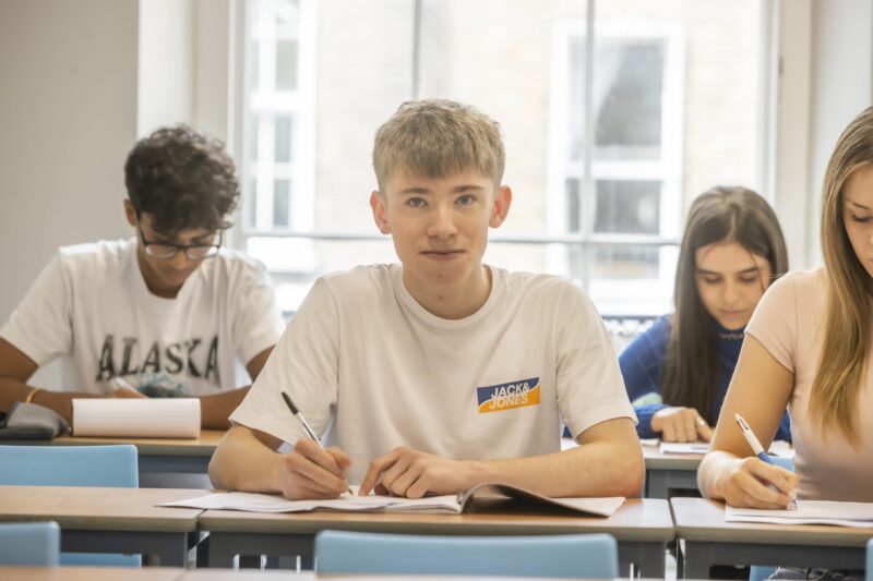 A student sitting at a desk in class looking towards the camera, with classmates writing in notebooks around him.