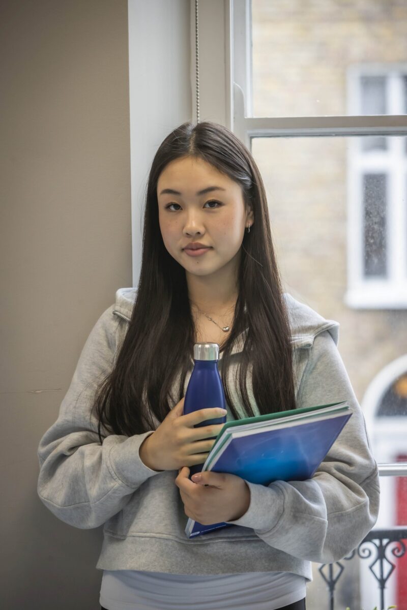 A student standing by a window, holding a water bottle and folders.