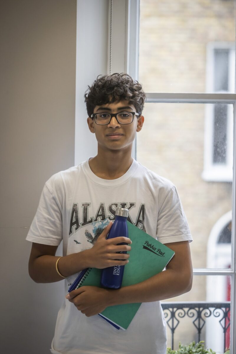 A student standing by a window, holding a water bottle and folders.