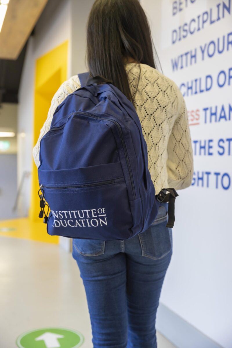A student walking down a corridor carrying an Institute of Education branded navy backpack, with a quote wall visible alongside her.