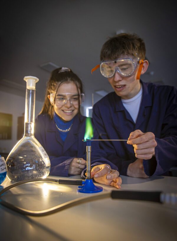 Two students in navy lab coats and safety goggles conducting a flame test experiment with a Bunsen burner and flask.
