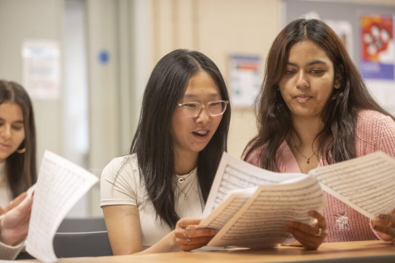 Two students reading sheet music together during a class, with a third student partially visible beside them.