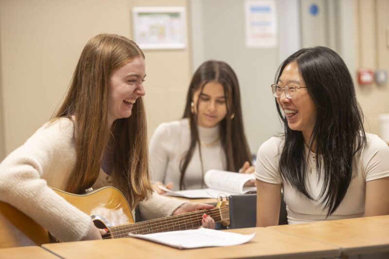 Three students laughing together, one playing guitar, in a relaxed classroom or common room setting.