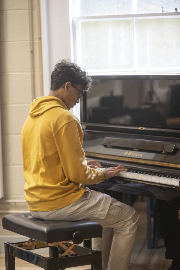 A student playing an upright piano in a practice room.