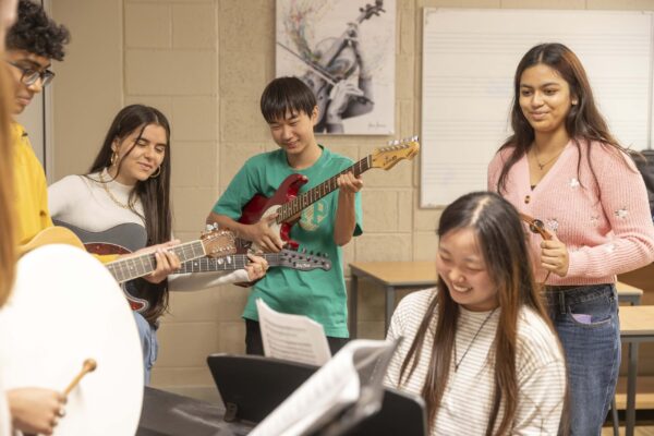 Five students play instruments and smile together in a classroom. One plays the piano, two have guitars, one holds a violin, and another plays a drum. Sheet music is on a stand in front of them.