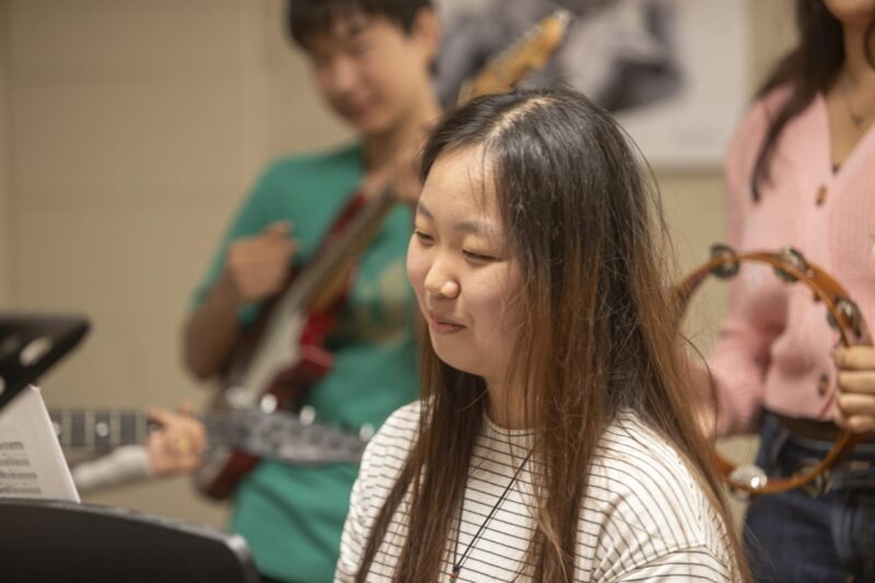 A student singing while classmates play guitar behind her during a music class.