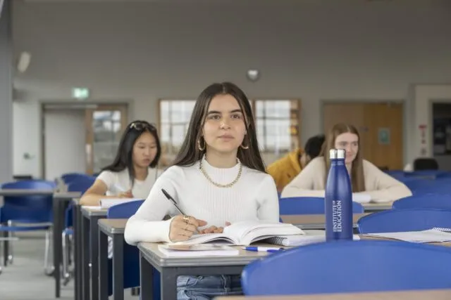 A young woman sits at a desk in a classroom, writing in a notebook with open books and a blue water bottle beside her. Other students are seated and working in the background.