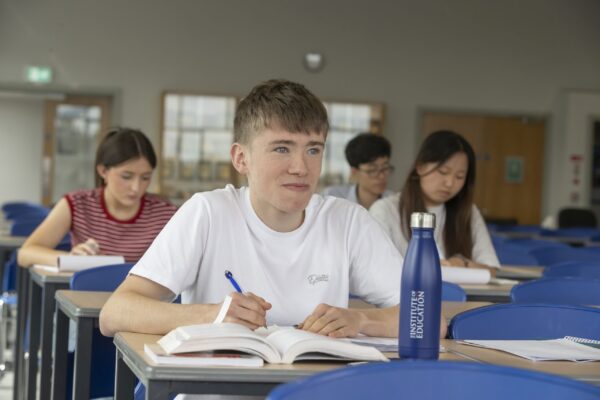 A student writing in a notebook at a desk during class, with classmates working at desks behind him in a bright classroom.