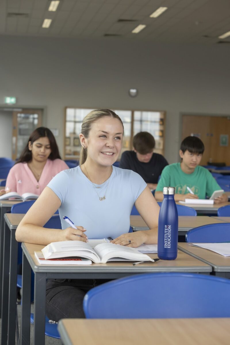 A smiling student sitting at a desk with an open notebook, an Institute of Education branded water bottle beside her, classmates seated behind in a classroom.