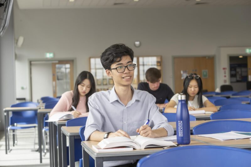 Male student sitting at a desk in a classroom, smiling at the camera with other students working in the background.