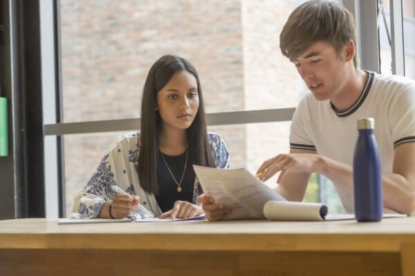 Two students sit at a table by a window, studying together. One points at notes whilst the other listens and writes. A blue water bottle sits on the table. They appear focused and engaged in their work.
