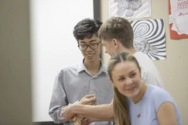 Three students looking closely together at something out of frame, in a classroom setting with artwork on the wall behind them.
