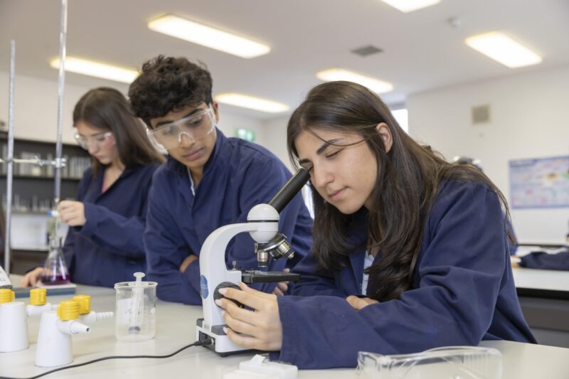 Three students in navy lab coats and safety goggles working with a microscope and lab equipment in a science lab.