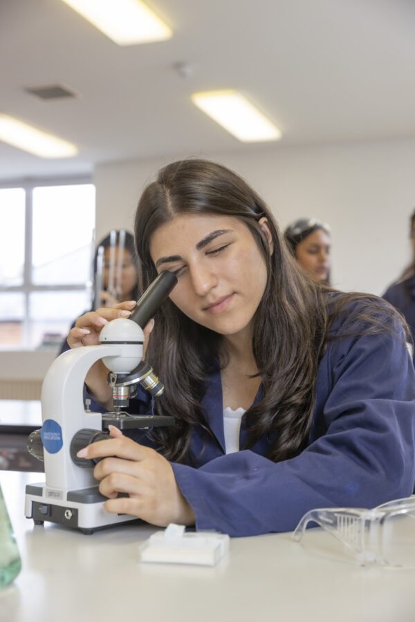 A student looking through a microscope at a workbench, with classmates in lab coats visible behind her.