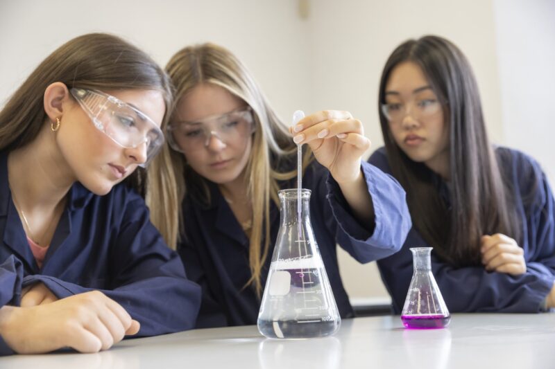 Three students in navy lab coats and safety goggles watching a colour-changing liquid being added to a flask during a chemistry experiment.