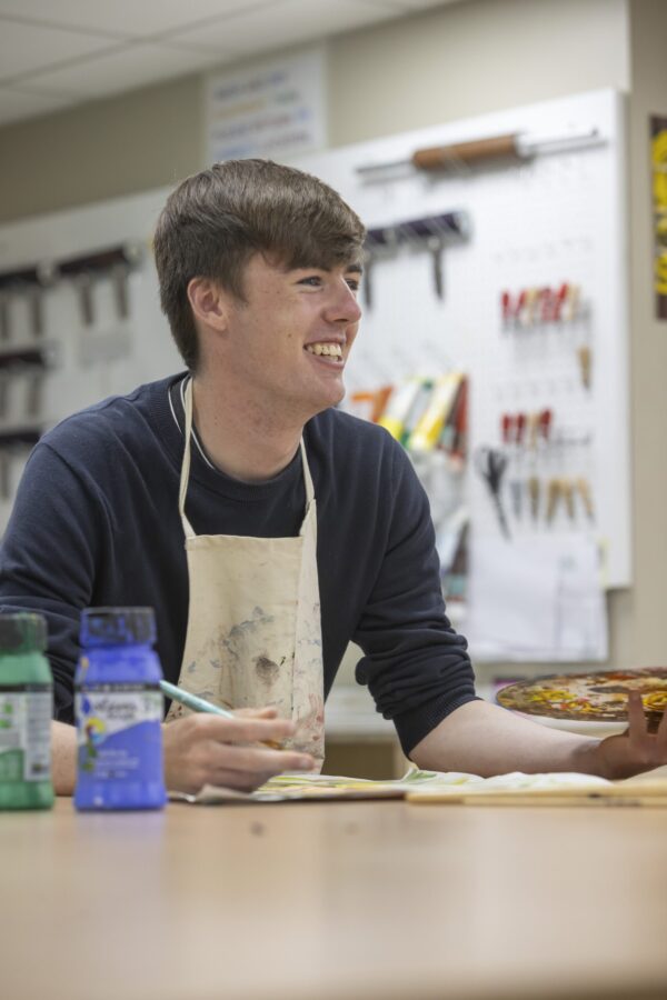 A student in an apron smiling while working at a table in a workshop or design classroom, with tools mounted on a pegboard behind him.