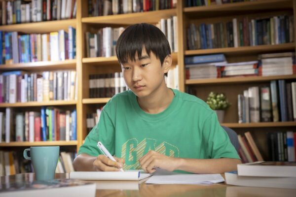 A student writing in a notebook at a desk, surrounded by tall bookshelves and stacks of books.