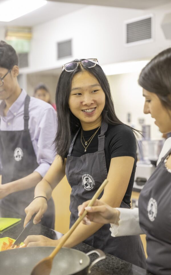 A smiling student in an Institute of Education branded apron taking part in a cooking class, with classmates working alongside her.